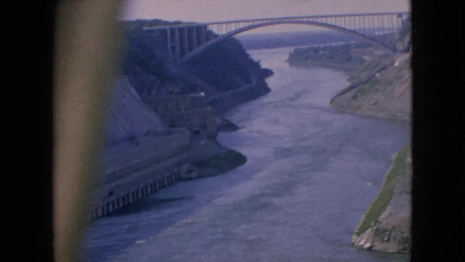 Vintage 8mm frame from the Niagara River area showing a high bridge above a broad river, with concrete works on the left bank and visible film-frame edges.