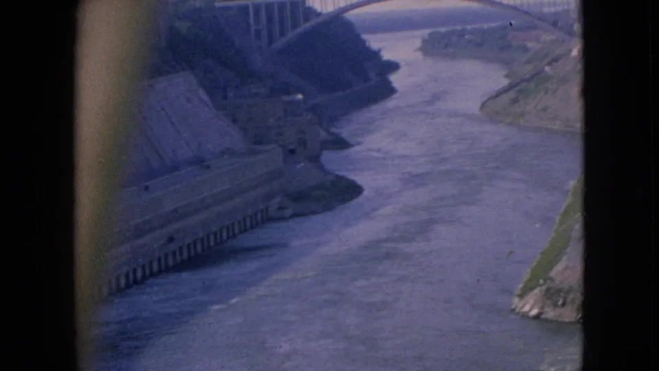 1964 home-movie frame looking across a Niagara-area river gorge toward a high arched bridge, with steep banks and dark borders from the filmed vantage point.