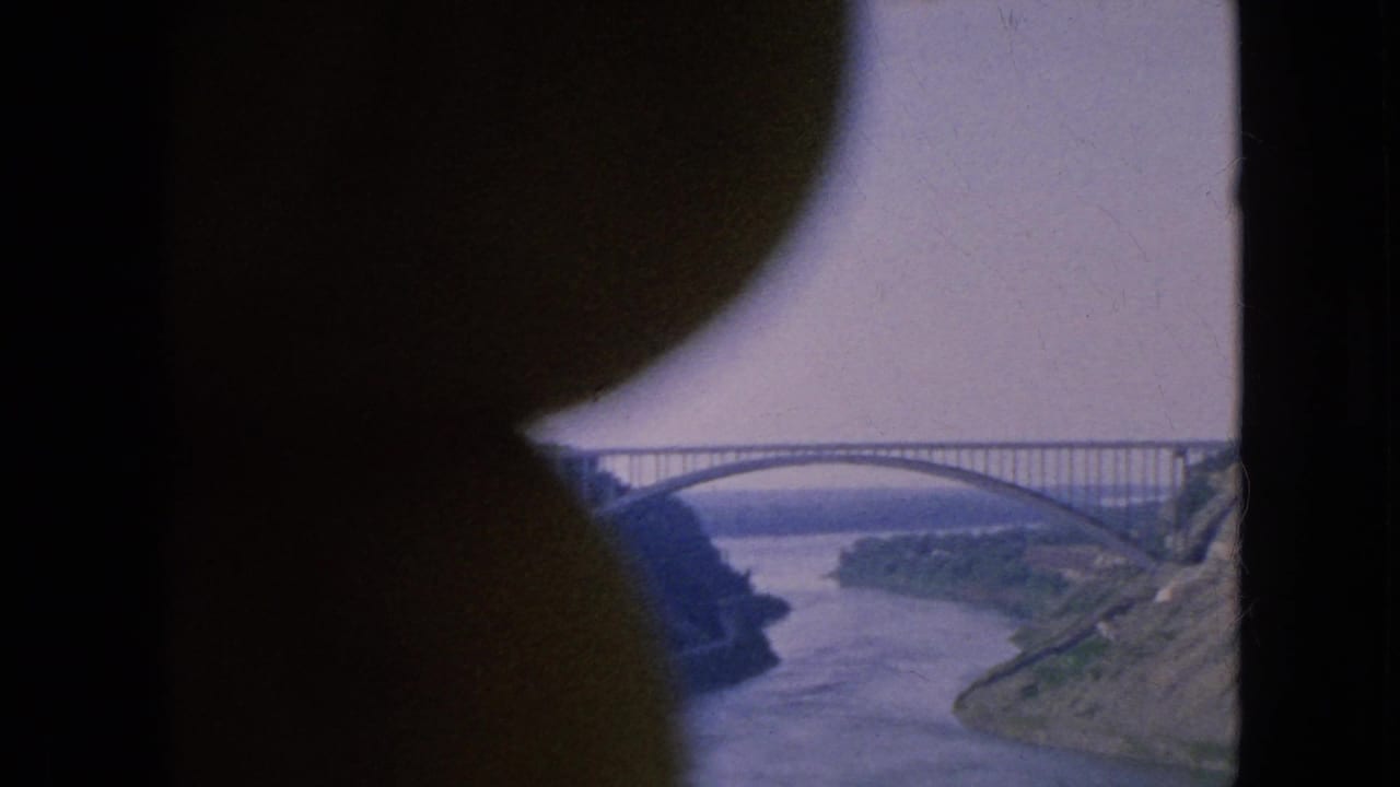 1964 Niagara Falls area home-movie frame showing a large arched bridge over a wide river, seen through a dark curved foreground obstruction.