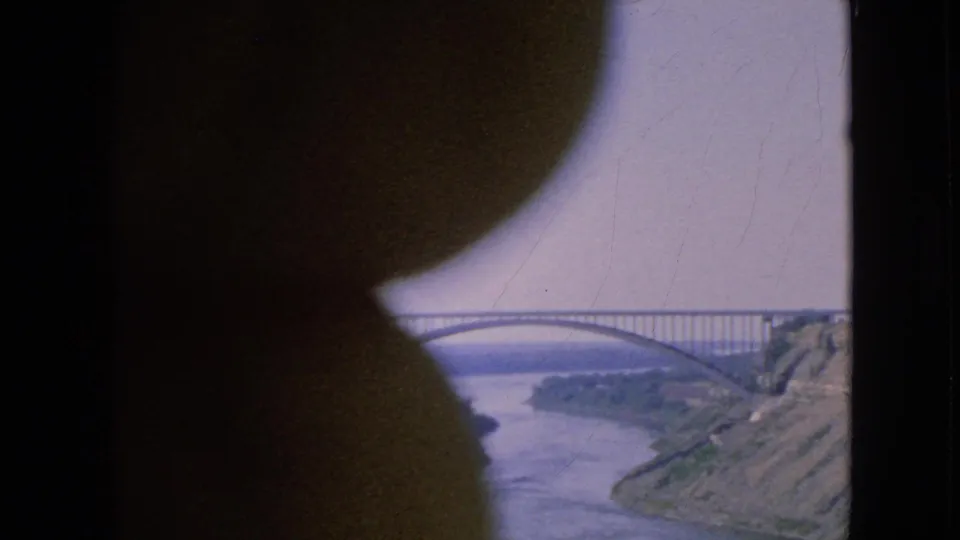 Vintage 8mm frame from 1964 showing a river, arched bridge, and sloping bank in the Niagara Falls area, with a dark rounded shape covering the left side.