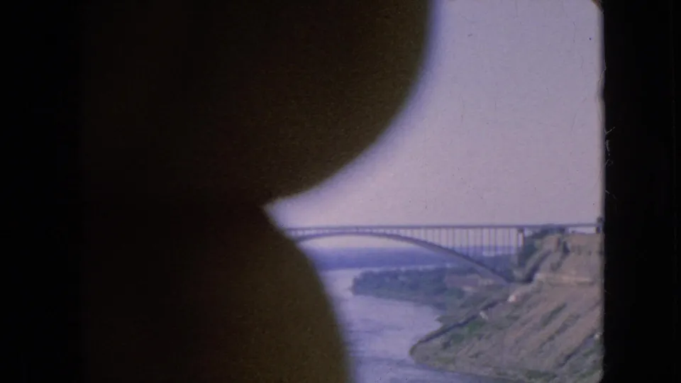 1964 Niagara Falls area travel footage frame showing an arched bridge over the river, rocky banks, and a dark curved foreground shape from the shooting position.