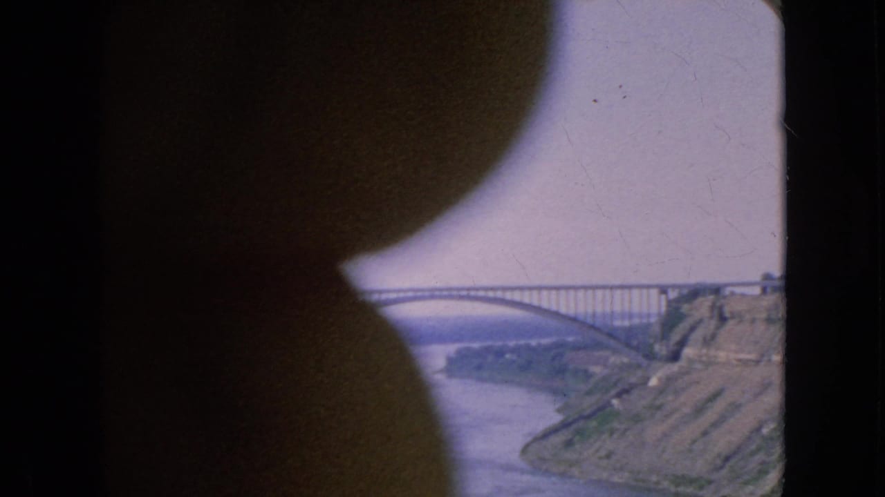 1964 home-movie view at Niagara Falls, New York, showing a steel arch bridge over water through a dark rounded foreground obstruction.