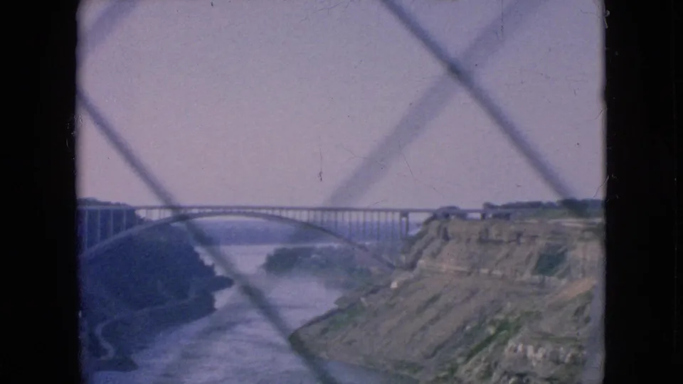 1964 Niagara Falls home-movie frame showing the bridge, river, rocky banks, and diagonal fence lines in the foreground.