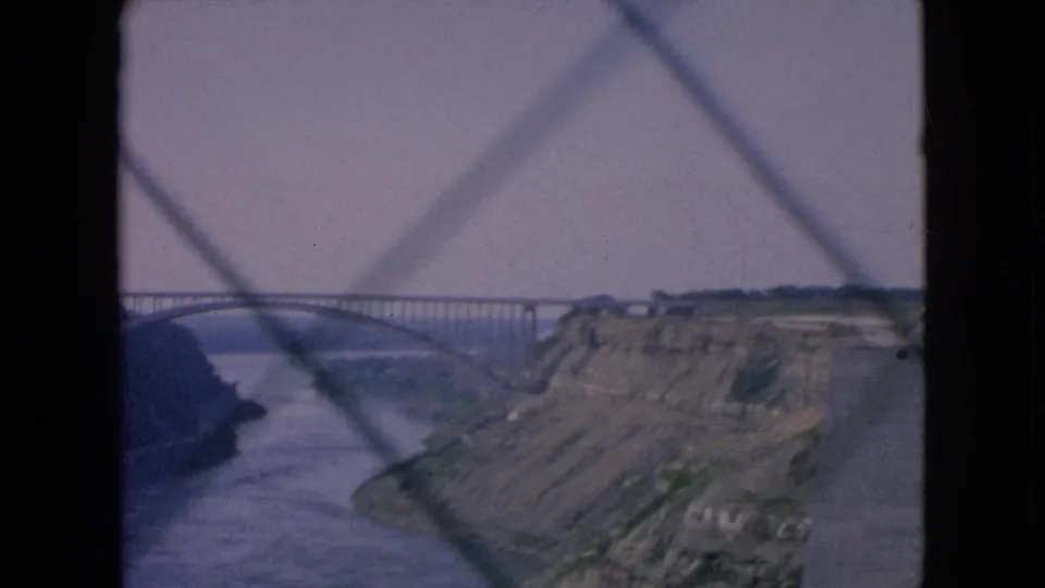 Soft 8mm frame of a steel bridge over the Niagara Falls river gorge, with blurred fence lines crossing the foreground.