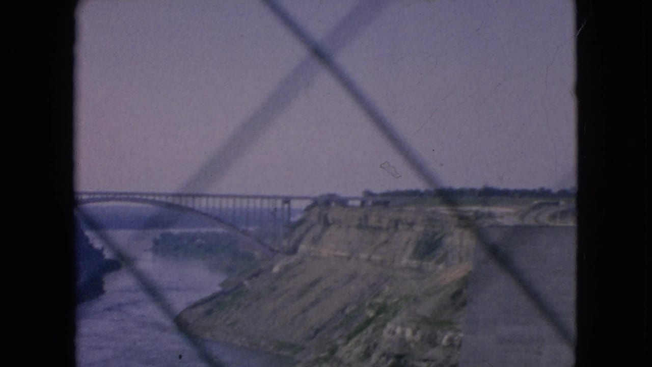 1964 8mm home-movie frame of a river gorge near Niagara Falls, New York, with an arched bridge and rocky cliffside viewed through a diamond-patterned barrier.