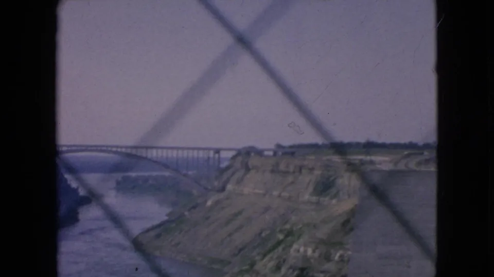 1964 8mm home-movie frame of a river gorge near Niagara Falls, New York, with an arched bridge and rocky cliffside viewed through a diamond-patterned barrier.