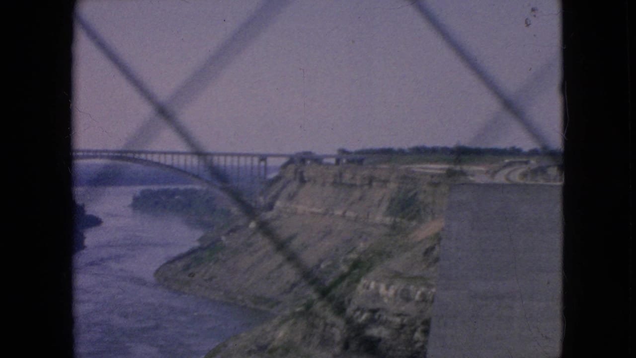 1964 Niagara Falls area travel footage frame showing a long arched bridge over a wide river gorge, with cliffs and a patterned foreground barrier in soft 8mm color.