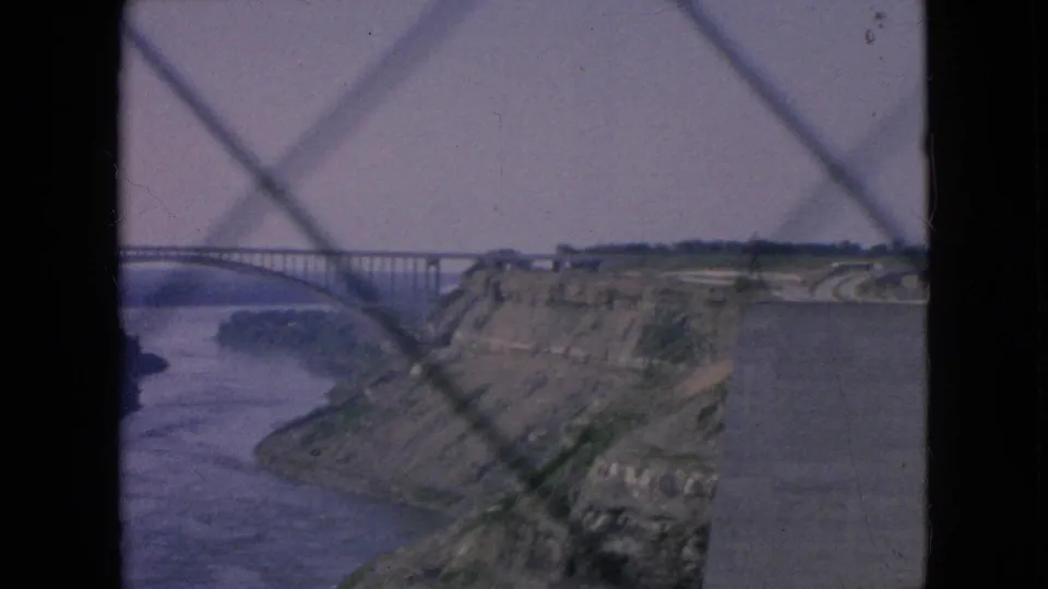 1964 Niagara Falls area travel footage frame showing a long arched bridge over a wide river gorge, with cliffs and a patterned foreground barrier in soft 8mm color.