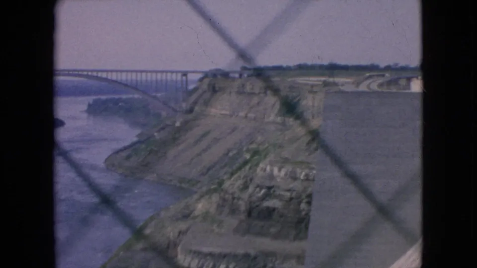 Home-movie frame from the 1964 Niagara Falls, New York clip showing the river, rocky gorge edge, bridge span, and curving roadway behind a dark diamond-patterned foreground.
