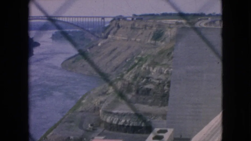 1964 8mm frame of the Niagara Falls area river gorge with an arched bridge, cliffside terrain, curving road, and a lower circular structure partly visible through the foreground grid.