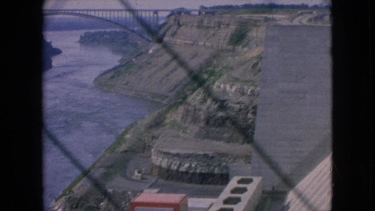 1964 8mm view of a Niagara Falls-area river gorge with concrete power-plant or dam structures, rocky slopes, and foreground window or fence lines.