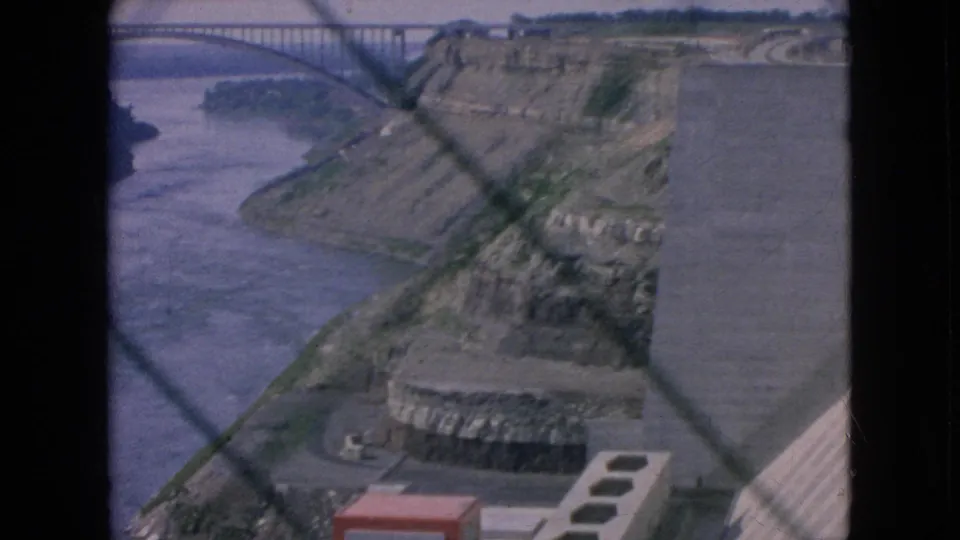 1964 8mm view of a Niagara Falls-area river gorge with concrete power-plant or dam structures, rocky slopes, and foreground window or fence lines.