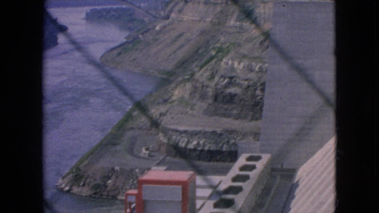 Concrete dam structure, rocky bank, river, and red utility box in foreground.