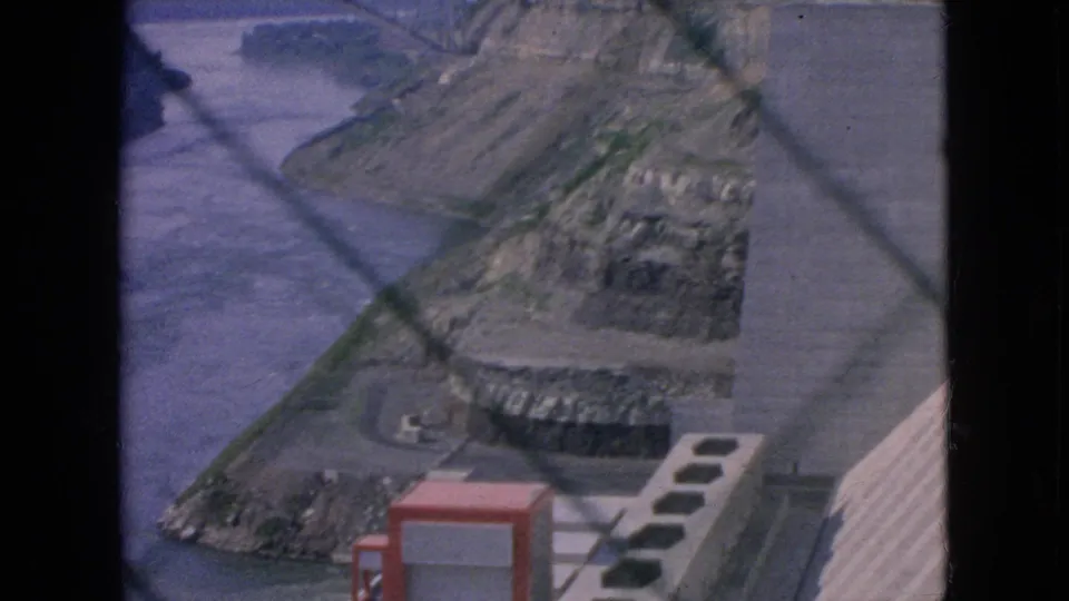 1964 8mm frame showing Niagara Falls-area hydroelectric infrastructure with a river channel, cut rock face, concrete works, paved service area, and red utility enclosure.