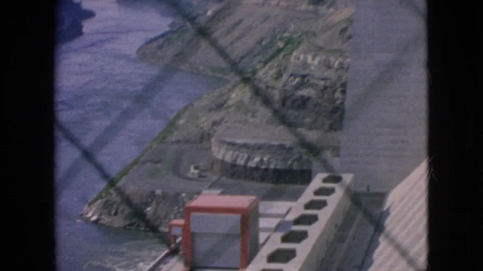 Visitor-shot 1964 8mm view of concrete walkways, a red utility enclosure, river water, and layered dam or power-plant structures at Niagara Falls, New York.