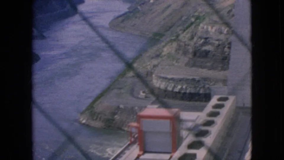 1964 8mm frame of the Niagara Falls-area river channel with concrete power infrastructure, rocky banks, service paving, and a red utility box partly in view.