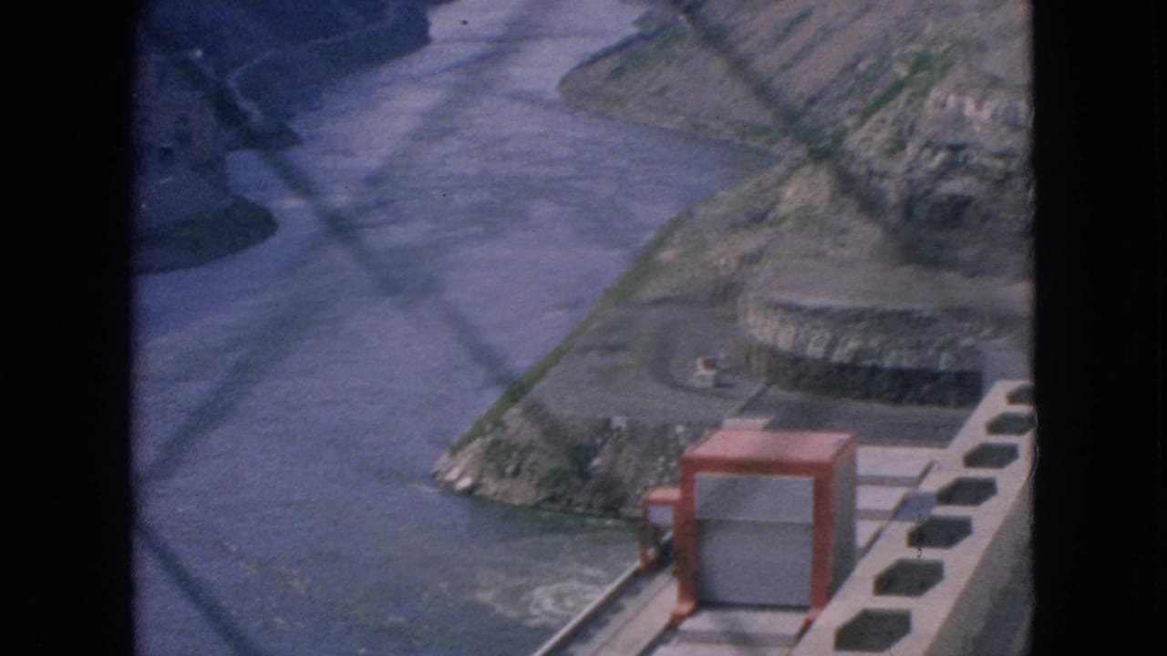 1964 8mm home-movie frame from Niagara Falls, New York, showing a river at the base of a gorge through chain-link fencing with concrete infrastructure and a red box-like fixture below.