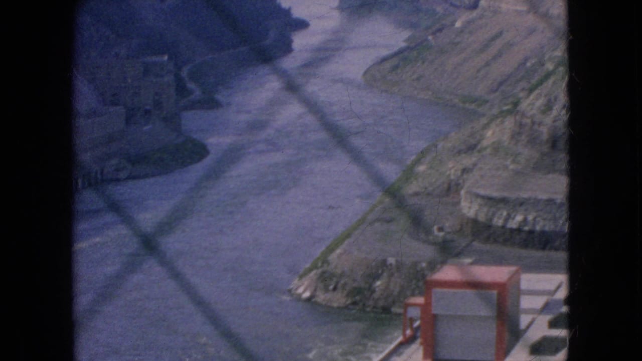 River and rocky gorge banks seen through chain-link fence with foreground concrete infrastructure partly visible.