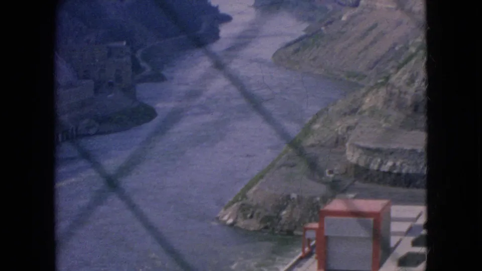 1964 Niagara Falls home-movie frame with the river running between steep gorge walls, seen through a chain-link fence from an elevated visitor vantage point.