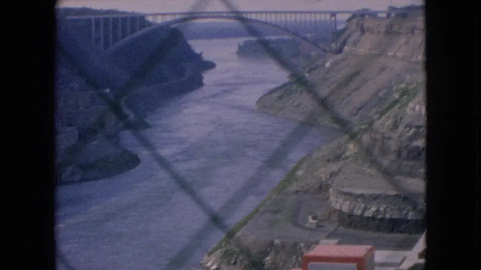 1964 8mm frame at Niagara Falls showing an arched bridge crossing the river gorge, with chain-link fence pattern over the view and concrete riverside structures below.