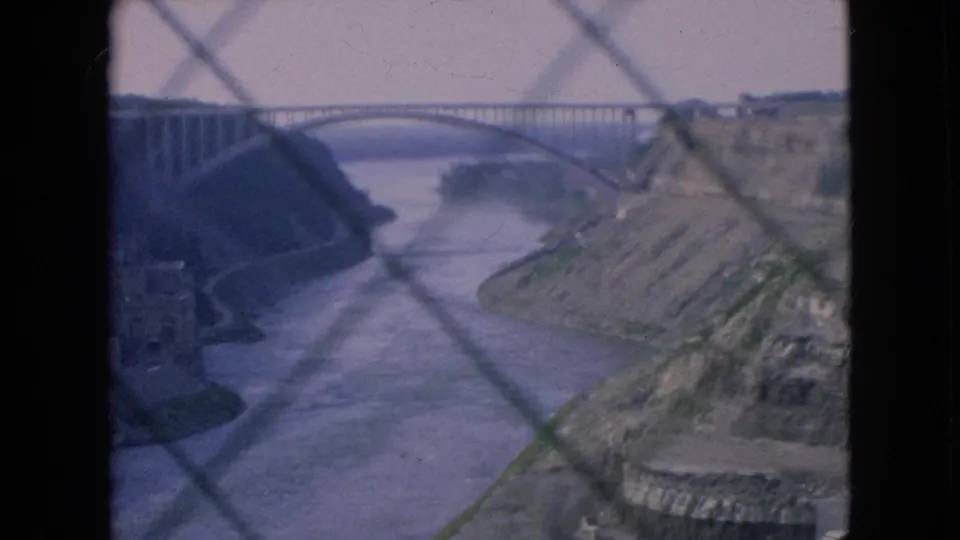 Niagara Falls, New York, 1964 home-movie frame of a large arched bridge spanning a river at the base of a gorge, filmed through foreground chain-link fencing.