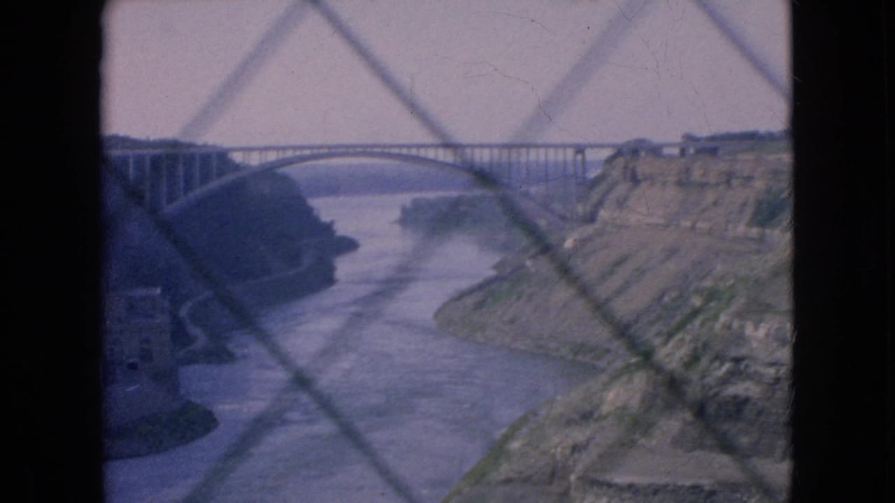 1964 Niagara Falls-area 8mm frame showing a steel arch bridge over a river gorge, viewed through a diamond-patterned screen or barrier.