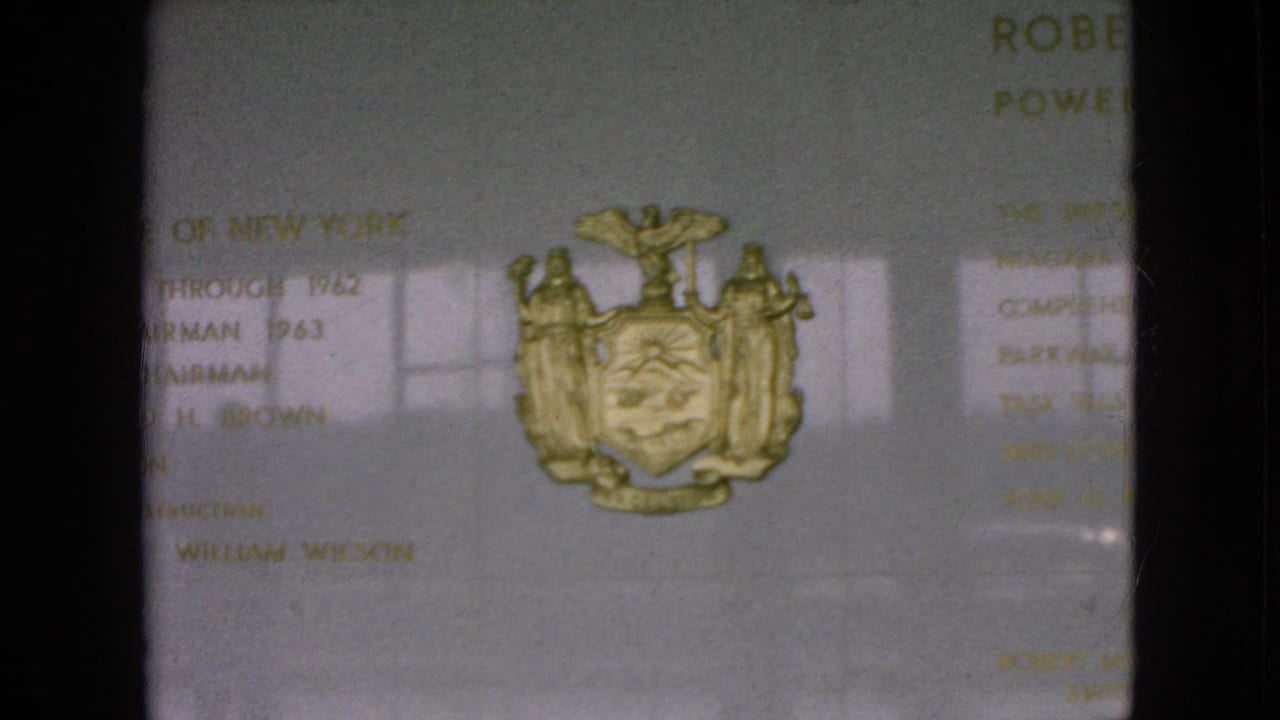 1964 Niagara Falls, New York home-movie frame showing a commemorative power authority plaque with a gold New York State coat of arms, soft lettering, and window reflections.