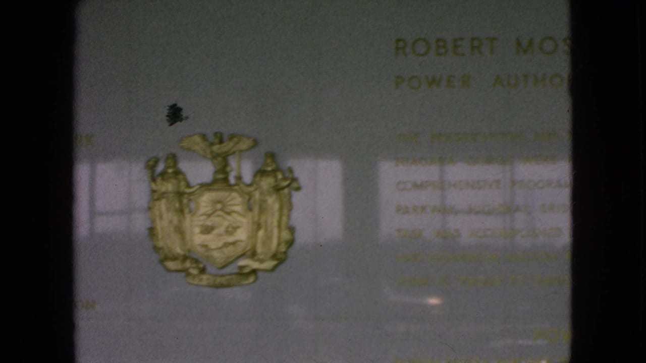 1964 Niagara Falls, New York frame showing a lower view of the commemorative plaque with the gold coat of arms, partial Robert Moses text, and visible film gate shadows.