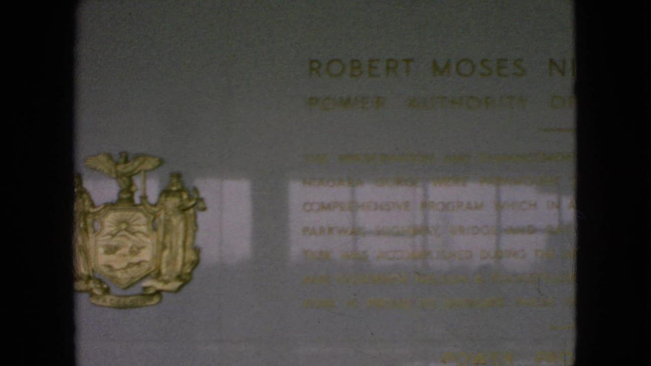 1964 8mm frame showing part of a Robert Moses Niagara Power Plant commemorative plaque on a light wall, with the New York State seal at left and soft reflected light across the surface.