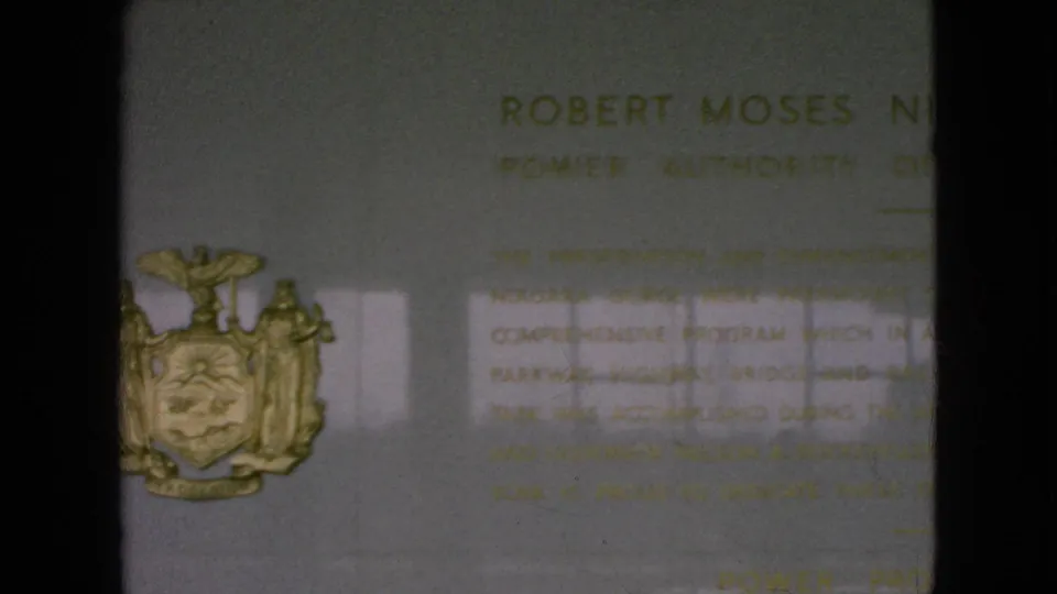 1964 8mm frame showing part of a Robert Moses Niagara Power Plant commemorative plaque on a light wall, with the New York State seal at left and soft reflected light across the surface.