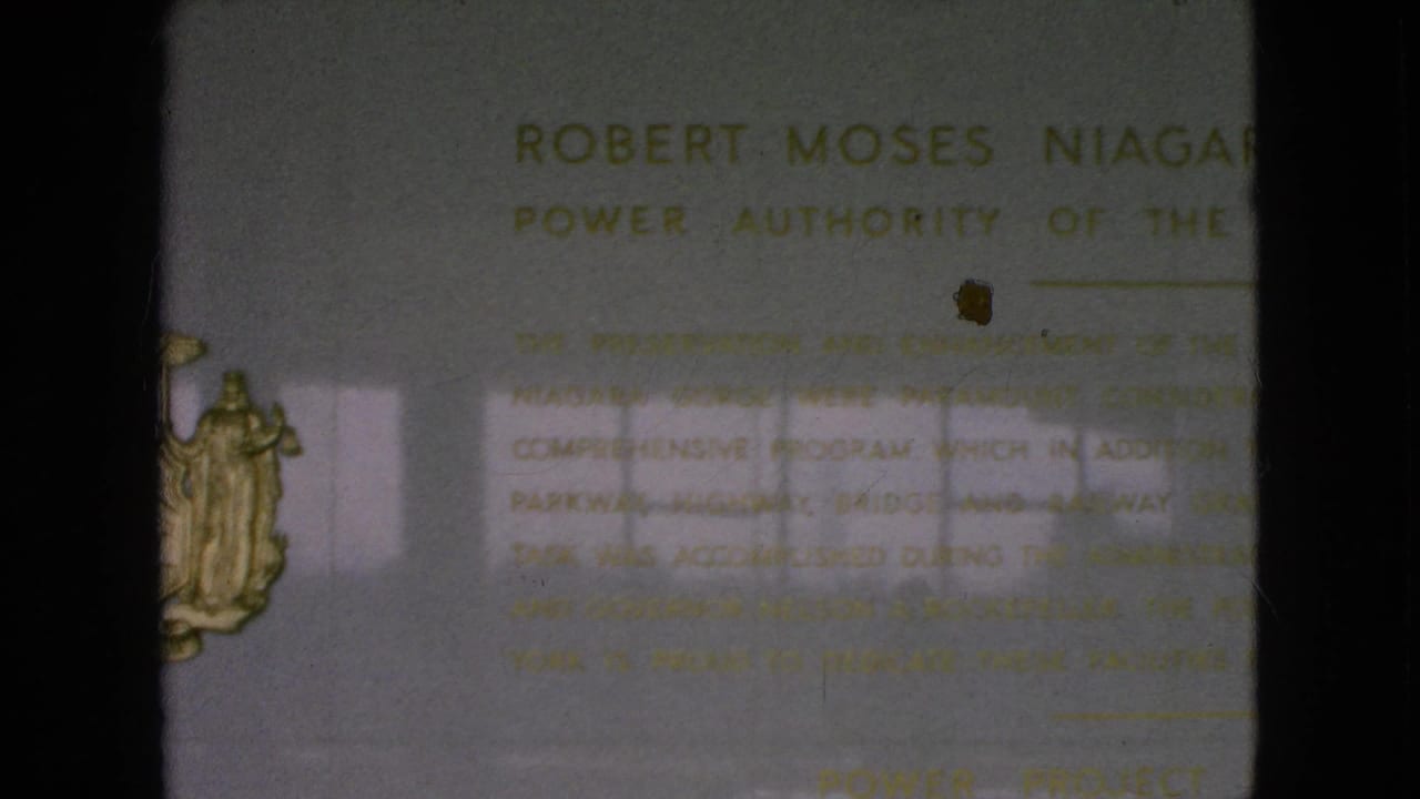 Close 8mm frame of the Robert Moses Niagara Power Plant plaque, showing gold inscription text, a partial New York State seal, and visible film grain on the pale wall surface.