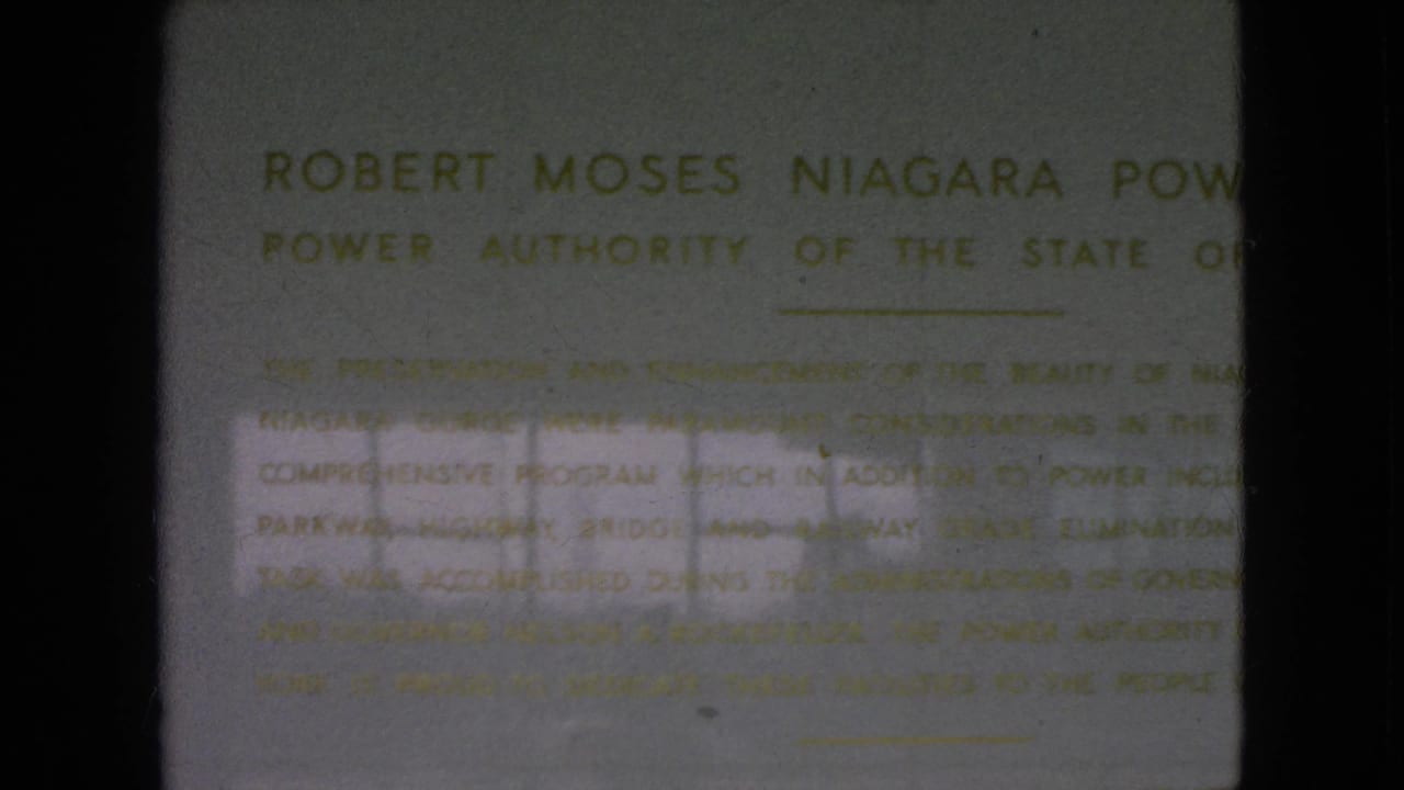 1964 home-movie frame holding on the lower portion of the Robert Moses Niagara Power Plant plaque, with gold text softened by focus and exposure variation.