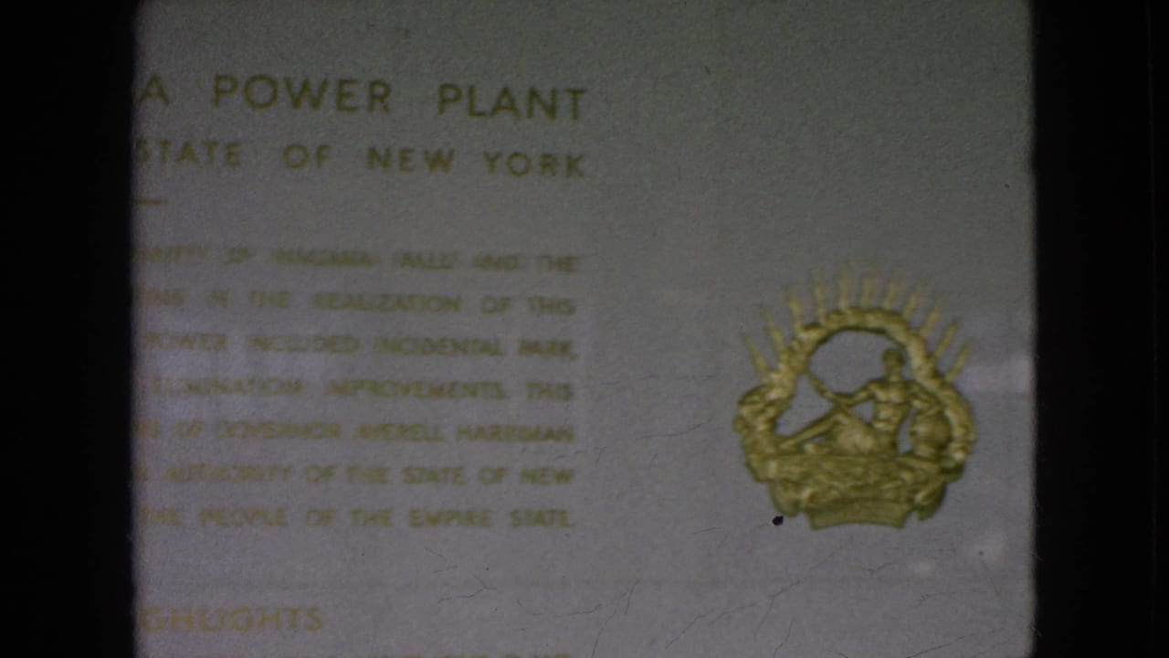 1964 8mm frame showing the lower area of the Moses Niagara Power Plant tablet, including blurred gold text and a decorative gold emblem.