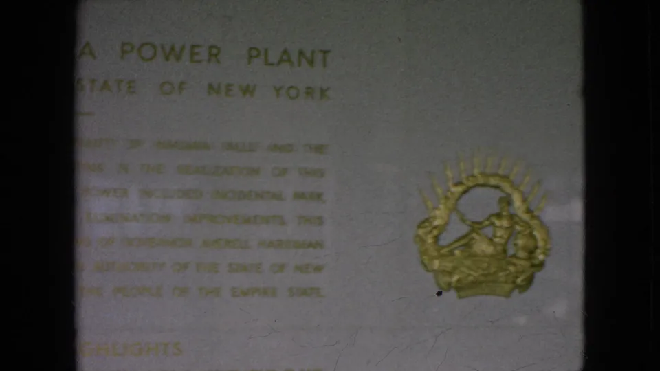 1964 8mm frame showing the lower area of the Moses Niagara Power Plant tablet, including blurred gold text and a decorative gold emblem.