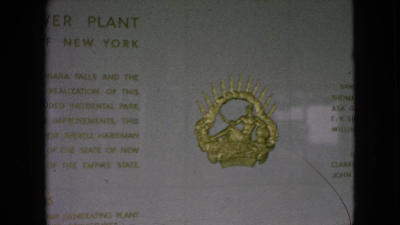 A close 1964 Niagara Falls, New York home-movie frame shows a plaque with partial text and a gold emblem resembling a merman or figure within a sunburst.