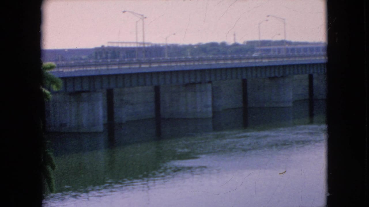 The home-movie frame continues the Niagara Falls bridge view, showing the span, concrete piers, roadway traffic, and rippled river surface below.