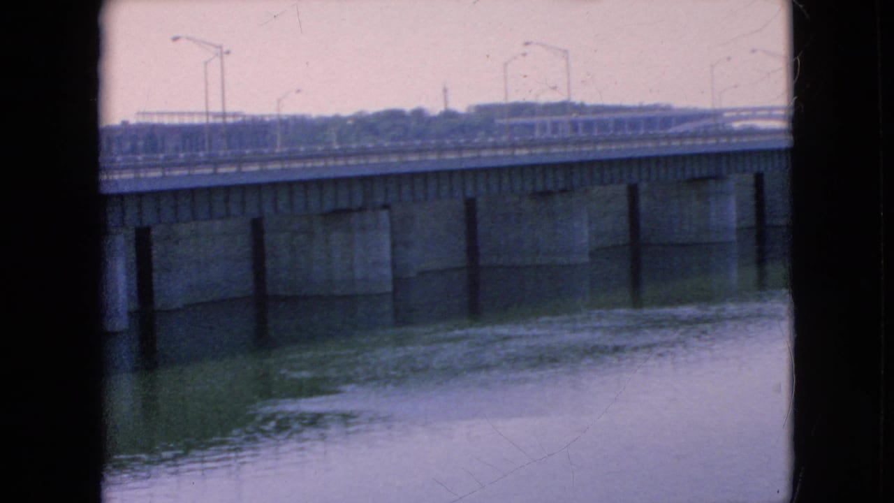 1964 8mm home-movie frame showing a concrete bridge over a wide river in the Niagara Falls, New York area, with streetlights along the roadway and a distant treeline under an overcast sky.