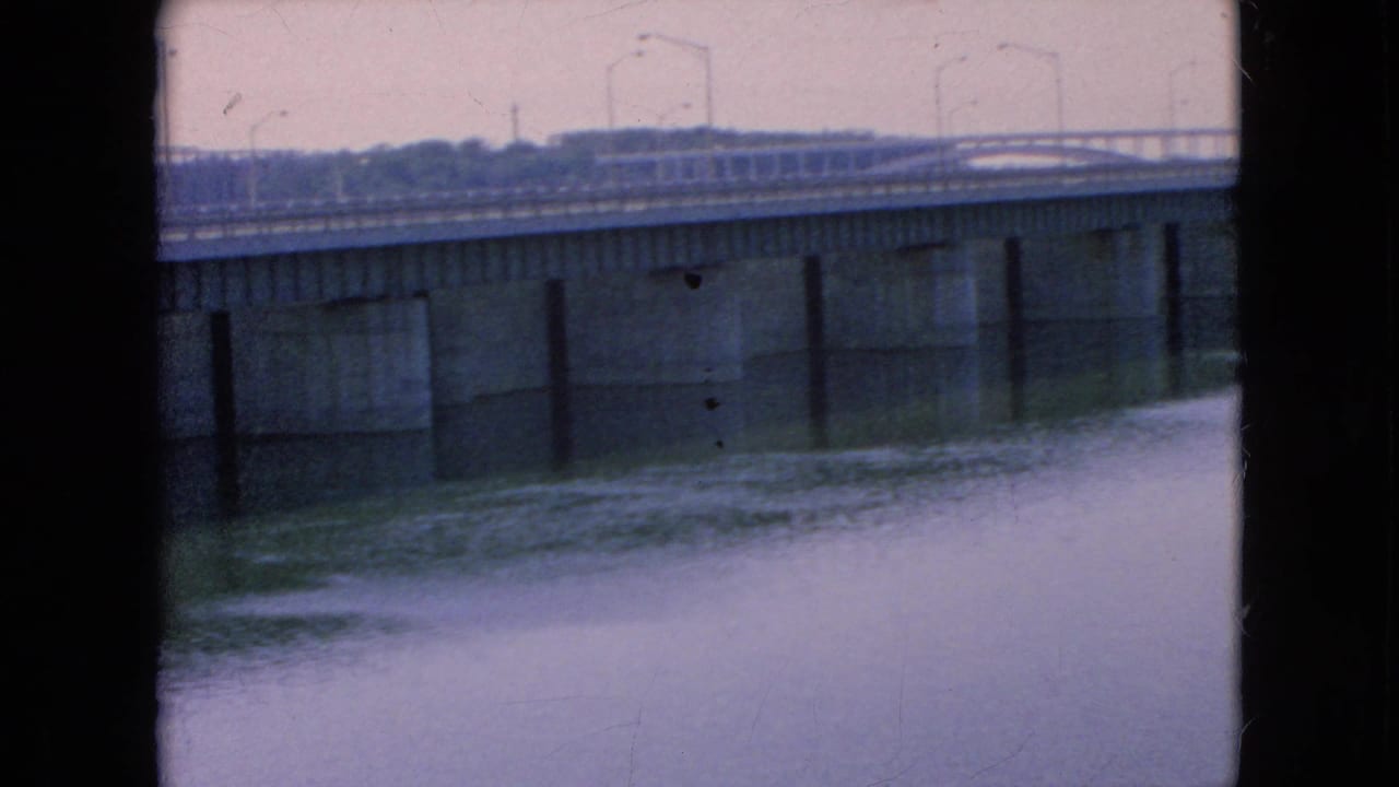 A soft 8mm frame of the bridge crossing the river, with repeated streetlights on the upper roadway, concrete supports below, and dark film borders visible at the sides.