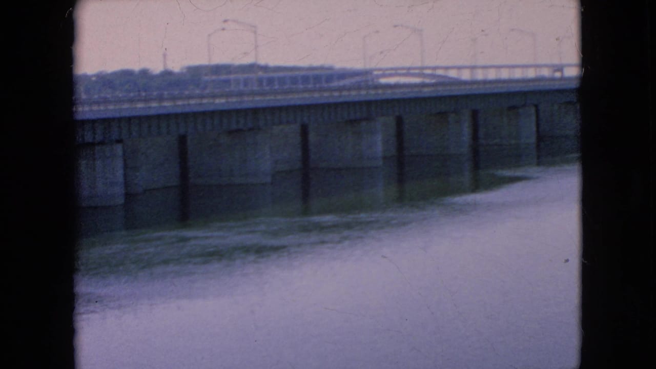1964-era home-movie view of a concrete river bridge where a small car appears on the roadway, with calm water below and the distant shoreline behind it.