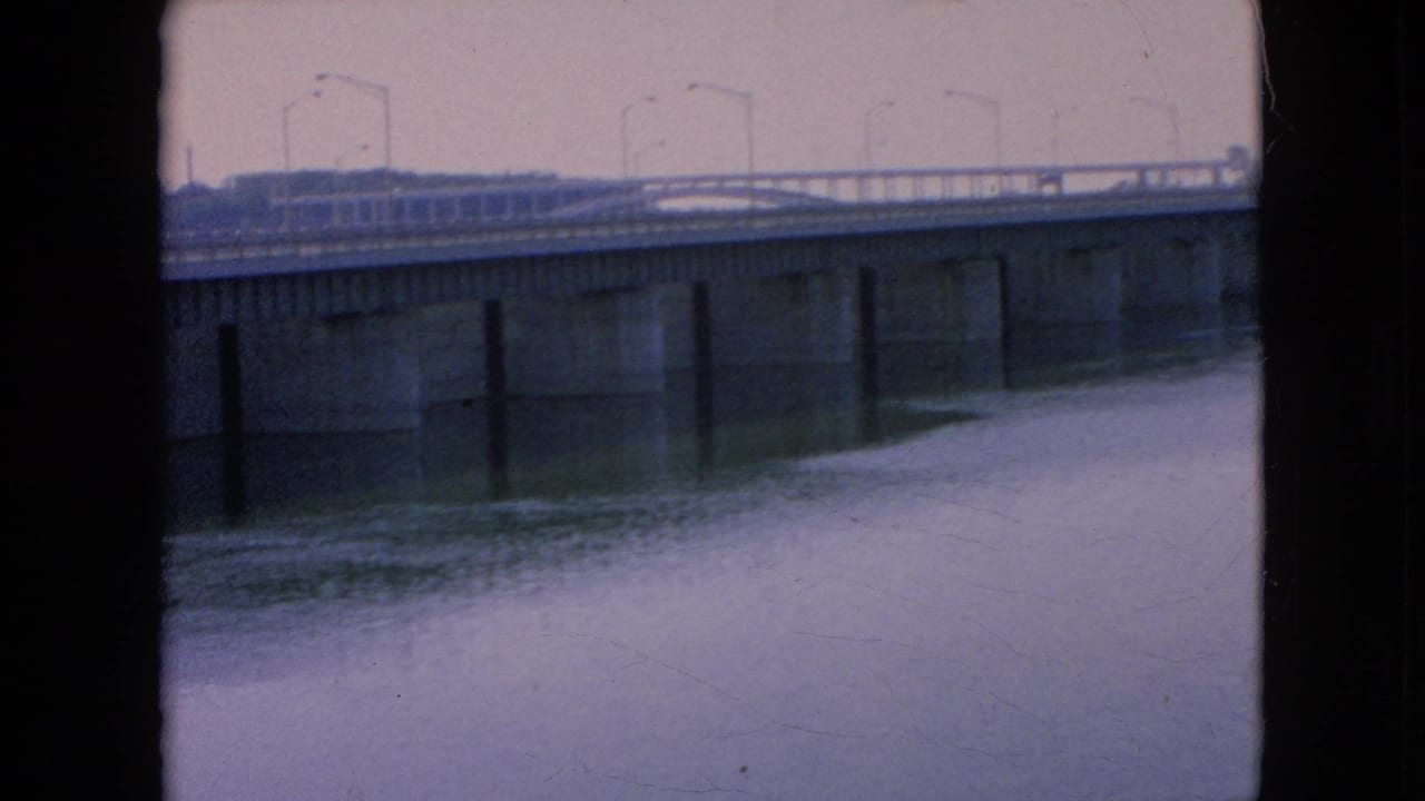 Final 8mm frame holds on the bridge spanning the river, with roadway lights silhouetted against the pale sky and the water filling the foreground.