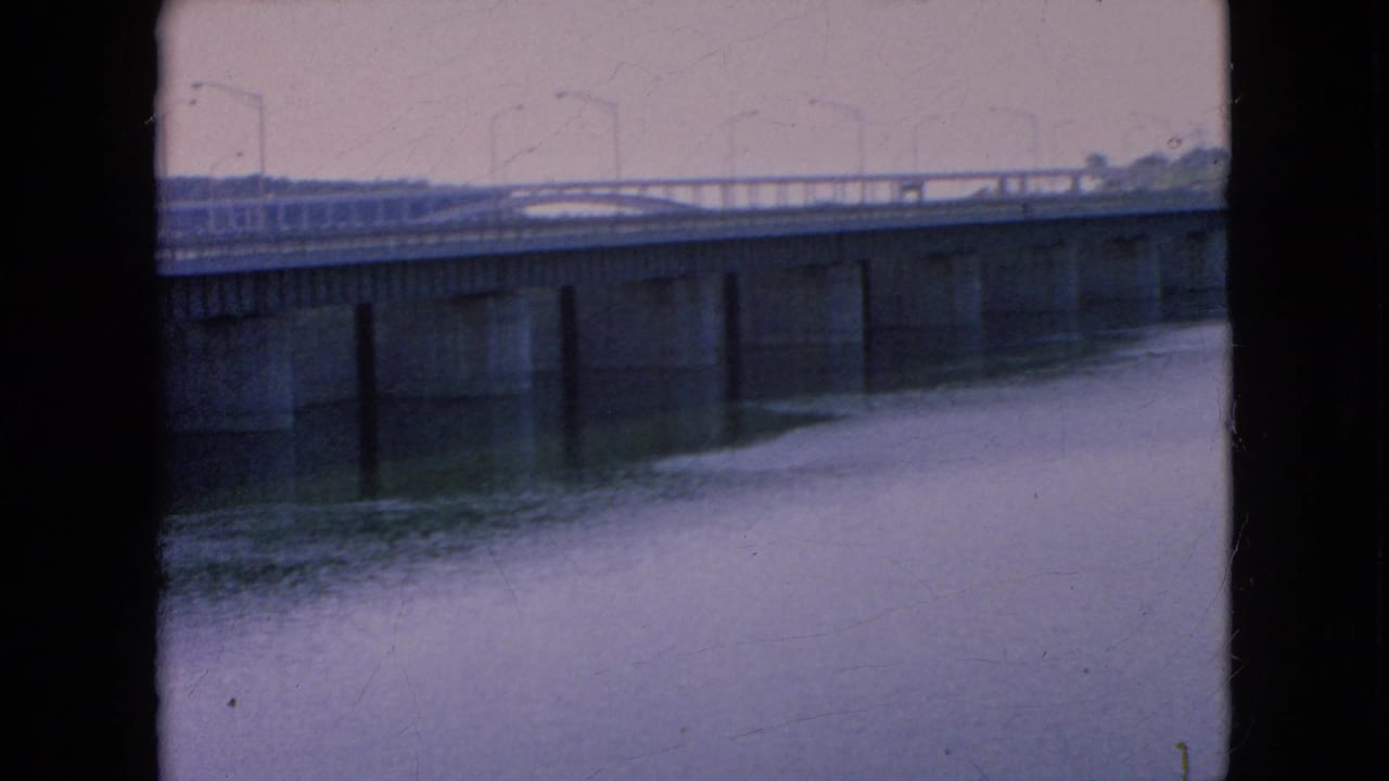 1964 8mm home-movie frame from the Niagara Falls, New York area showing a concrete bridge over still water beneath a muted grey sky.