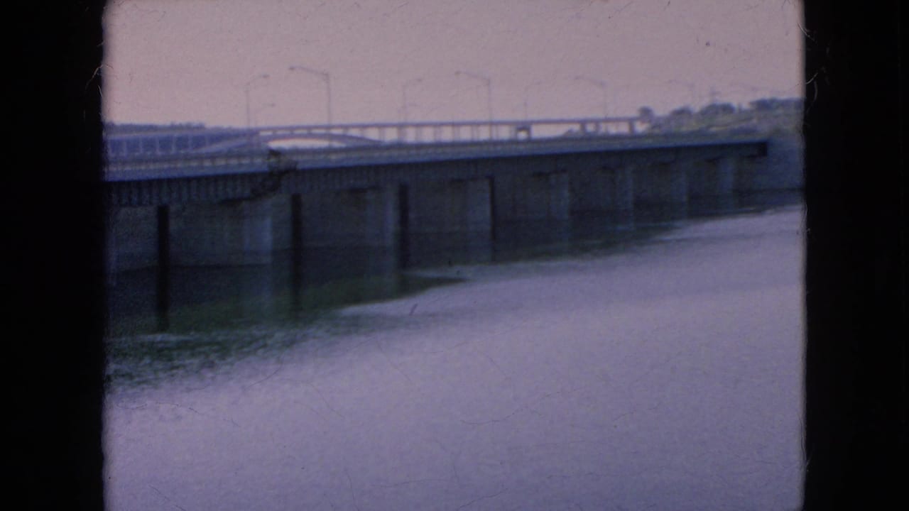 1964 Niagara Falls area home-movie view of a long bridge, concrete supports, calm water, and roadside lighting under cloudy conditions.