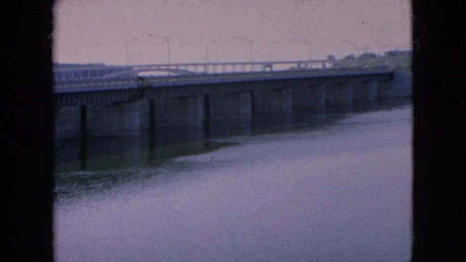 1964 Niagara Falls area home-movie view of a long bridge, concrete supports, calm water, and roadside lighting under cloudy conditions.