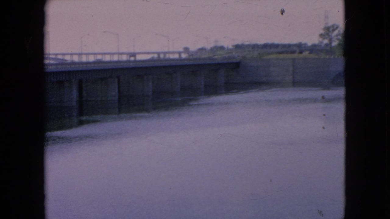 1964 8mm view in Niagara Falls, New York, showing a concrete bridge lined with lamp posts over a broad calm waterway, with soft focus and dark frame borders.