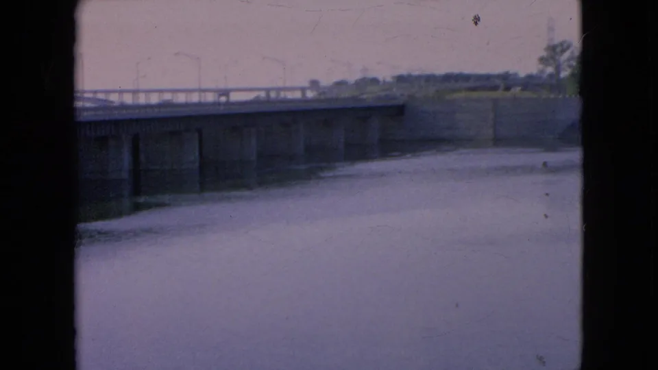 1964 8mm view in Niagara Falls, New York, showing a concrete bridge lined with lamp posts over a broad calm waterway, with soft focus and dark frame borders.