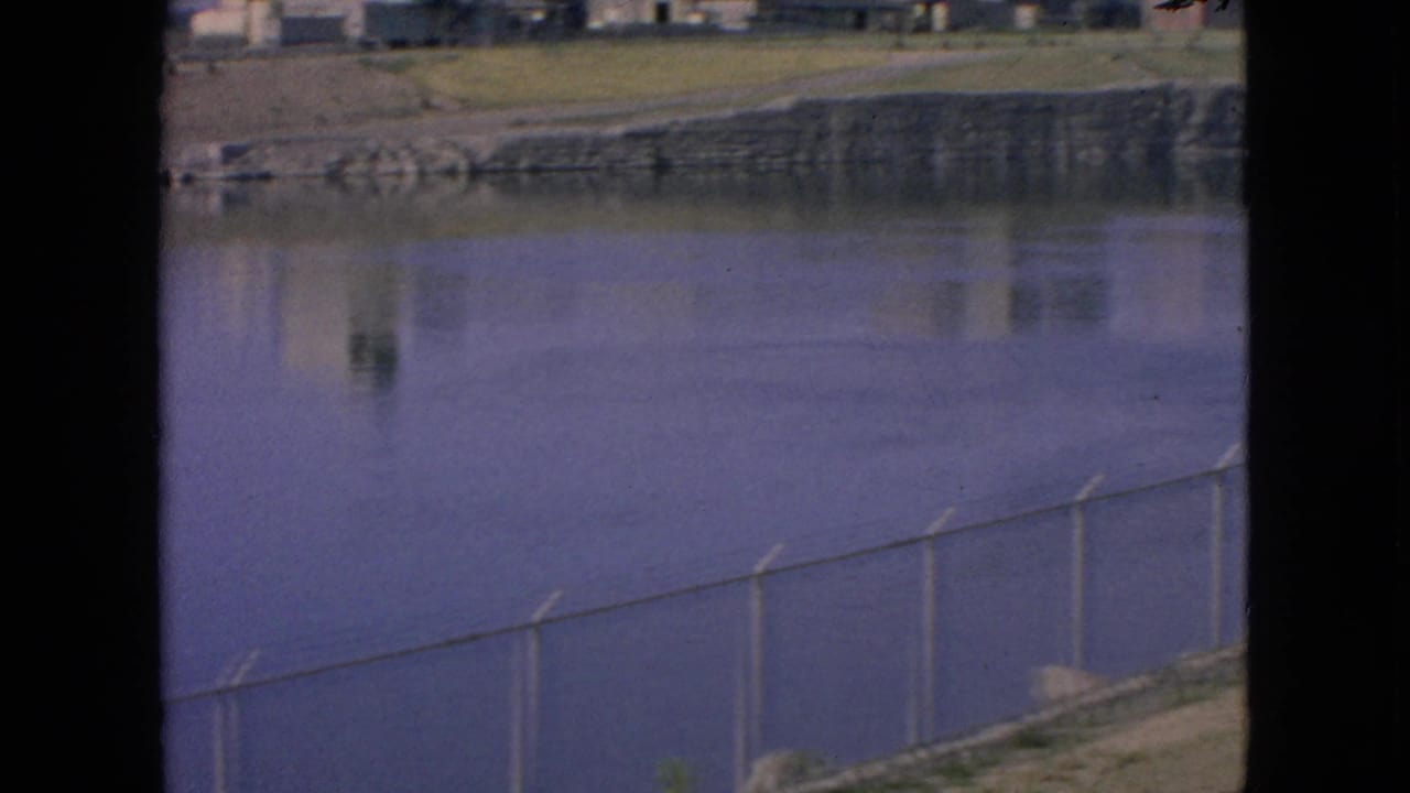 The final frame looks across the calm water toward a stone or concrete shoreline, with a chain-link fence in the foreground and faint building reflections on the surface.