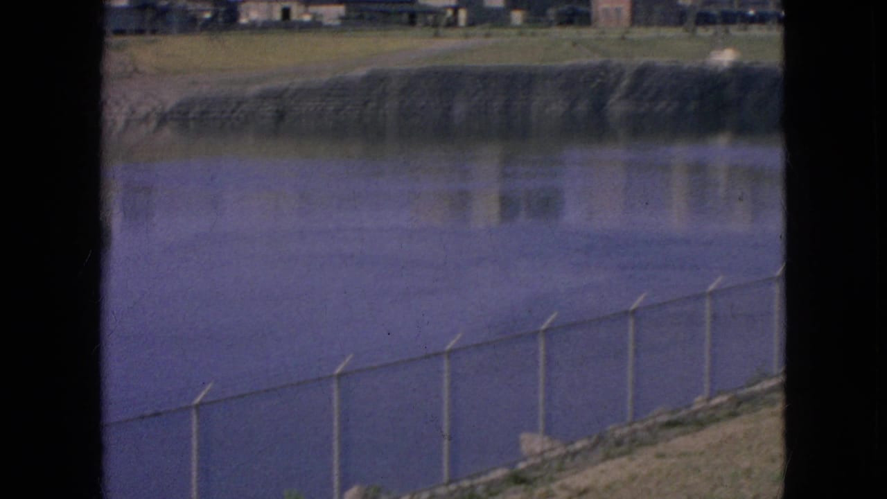 1964 Niagara Falls, New York 8mm frame showing calm water, a fenced shoreline, and industrial buildings across the river through dark window-like borders.