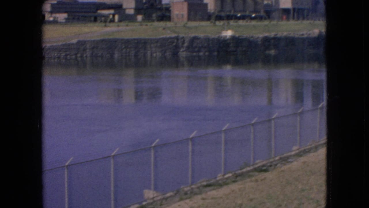 1964 Niagara Falls, New York frame with a chain-link fence running along the near riverbank and factory structures reflected in the water beyond it.
