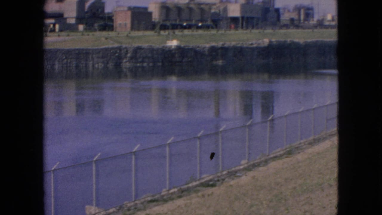 1964 Niagara Falls, New York home-movie frame showing a fenced shoreline in the foreground with low industrial buildings across calm water.