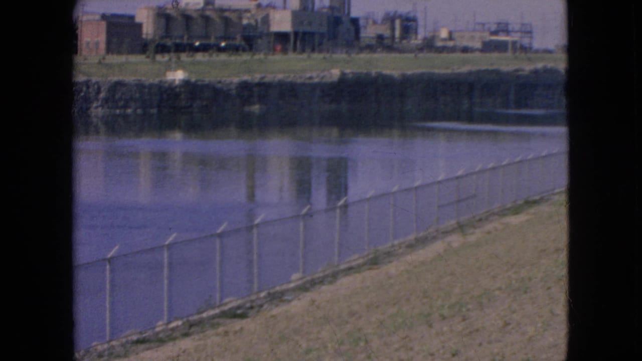 1964 Niagara Falls, New York frame showing a diagonal fence along the riverbank with industrial buildings and reflections across the water under clear light.